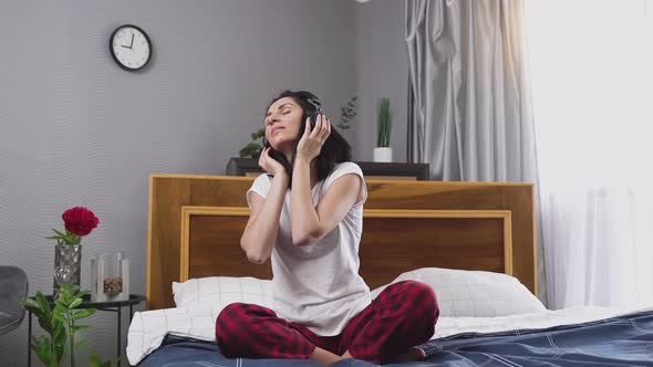 brunette in headphones sitting on bedroom bed and enjoying modern music and moving in the beat