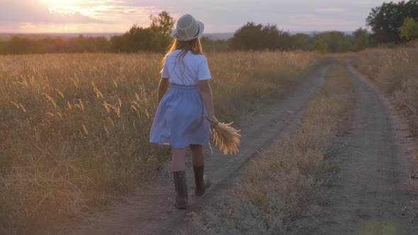 A Girl Walks Along a Rural Road at Sunset, Child in a Hat and Rubber Boots
