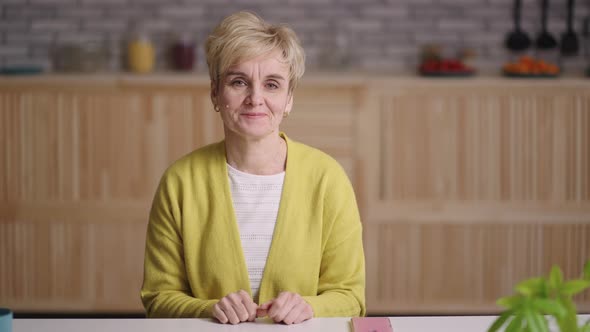 Portrait of Charming Old Lady Looking at Camera and Waving Hand to Welcome Sitting Alone at Kitchen