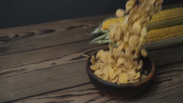 cornflakes fall into a bowl on a wooden table. ears of corn lie nearby.