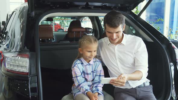 A Young Father with a Daughter Makes Selfie on the Background of a Car