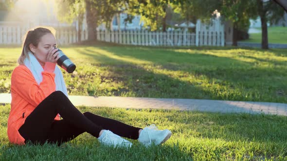 Young Beautiful Woman in a Red Sports Sweater Is Sitting on the Grass After a Morning Run.