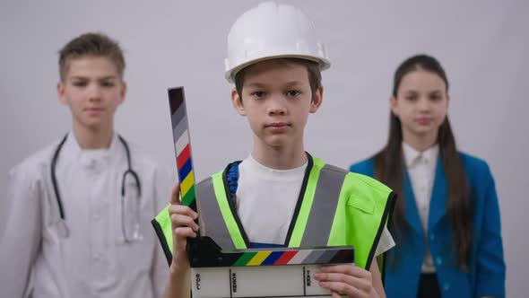 Unsure Boy in Hard Hat and Builder Uniform Standing with Clapper Board Looking at Camera As Friends