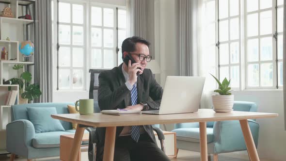 Asian Businessman Answering The Phone While Using The Computer For Working At Home