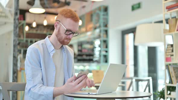 Redhead Man Using Smartphone and Laptop in Cafe 