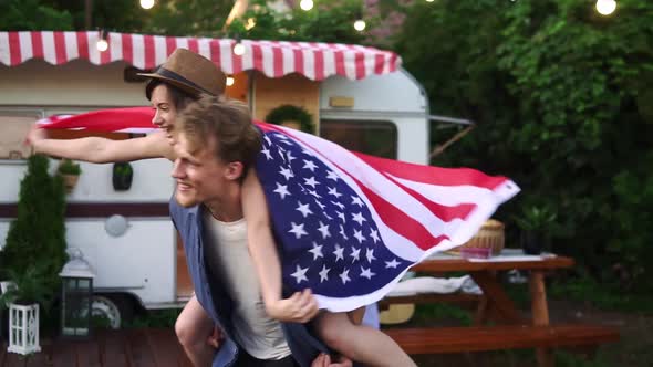 Carefree Cheerful Couple Having Fun Near Trailer in the Park Man Piggybacking Girl with American