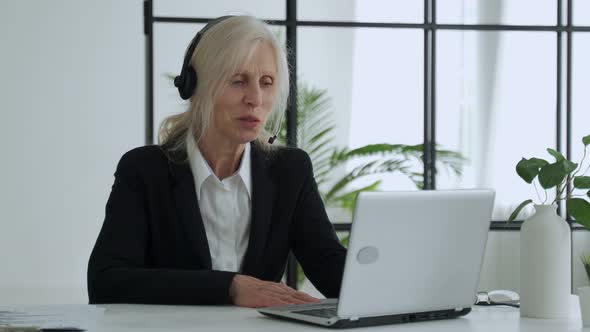 Elderly Lady in a Headset Talks on a Video Conference Conducts a Video Conference Using a Laptop in