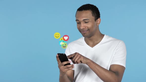 African Man Using Smartphone Isolated on Blue Background Flying Smileys Emojis and Likes