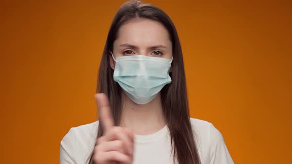 Pretty Woman in Medical Mask Does Disapproving Gesture with Her Finger Against Yellow Background