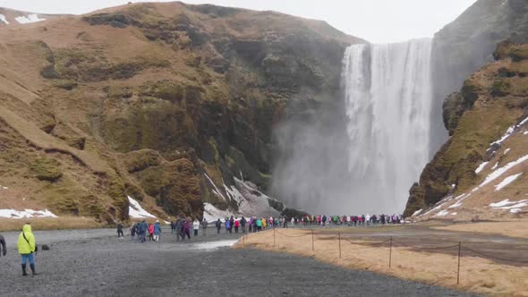 Static, slow motion shot of lots of people enjoying the view of Skogafoss waterfall, on a cloudy, au