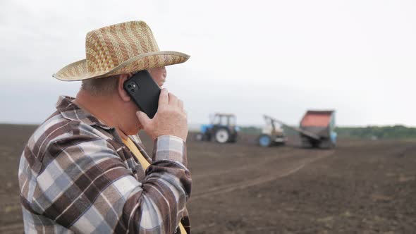 Senior Agricultural Businessman in a Hat Speaking on the Phone and Watching the Work of Combines in