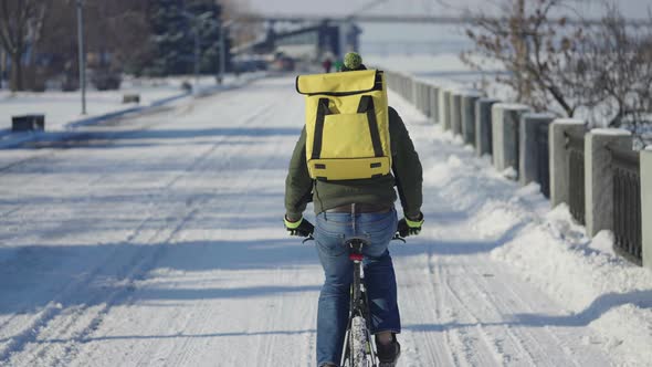 Back View of Man with Large Yellow Thermo Bag on His Back Rides Bicycle Along Snowy Street and