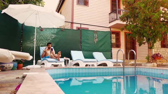 Man Lies on a Lounge Chair in Front of Pool Side and Talks By Video Call Using Smartphone in His