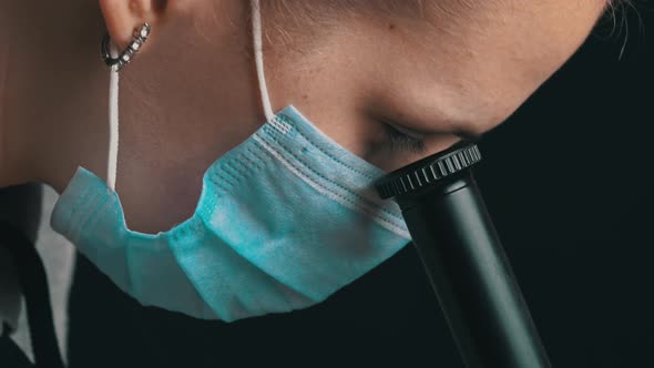 Girl Scientist Looks Into the Lens of a Microscope in a Mask Closeup