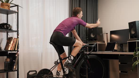 Male athlete is taking selfies on smartphone while doing cardio exercises on stationary bicycle
