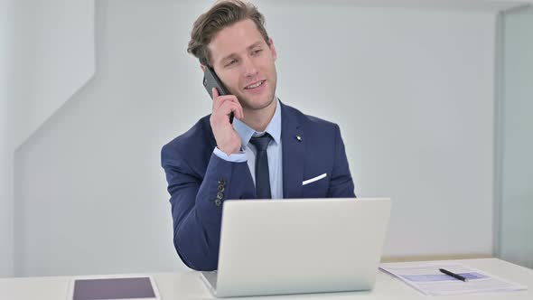 Serious Young Businessman Working on Laptop and Talking on Smartphone