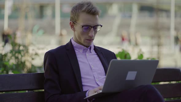 Concentrated Young Man Sitting on Bench and Typing on Laptop