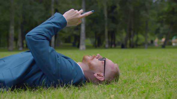 Confident Young Businessman Checking Phone Lying on Green Grass Outdoors