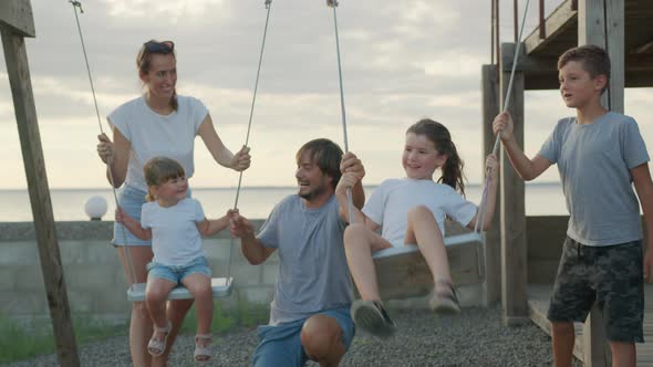 Happy Family Rolls Young Children on a Swing Outdoors at Sunset.