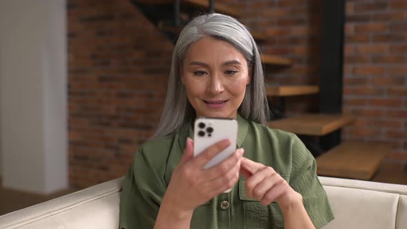 Surprised Senior Woman Looking at the Phone Screen Sitting on the Coach at Home