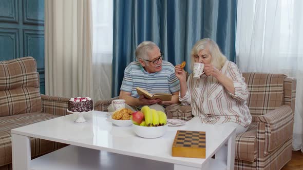 Retired Senior Couple Talking Drinking Tea Reading Book in Modern Living Home Room Lounge Together