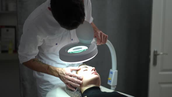 Focused Expert Male Beautician with Magnifying Lamp Examining Face of Female Client Indoors