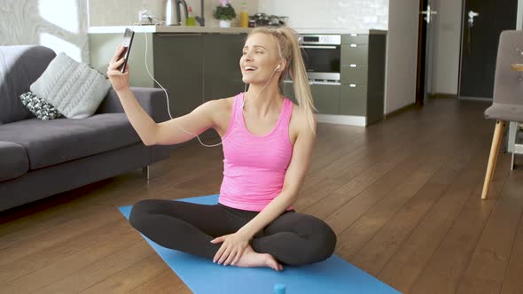 Young Attractive Sporty Woman Sitting on Yoga Mat Making Selfie with Her Mobile Phone