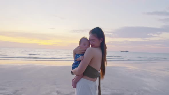 Happy mother with baby boy walks by ocean on the beach in summer