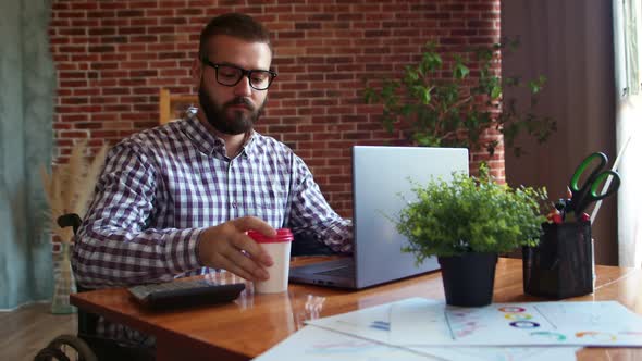 Man Wheelchair User is Working on Laptop Drinking Coffee Looking Ahead and Smiling