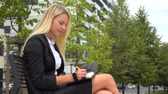 A Young Beautiful Businesswoman Sits on a Bench and Writes Notes in a Street in an Urban Area