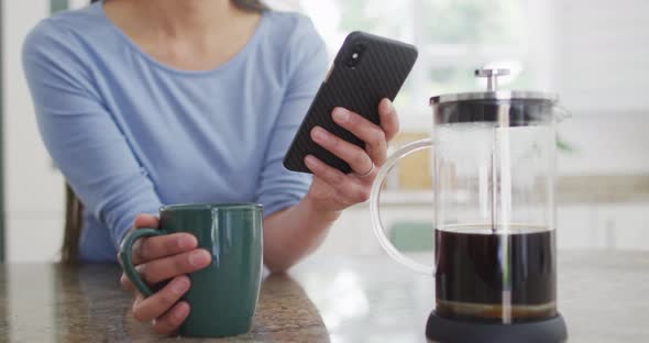 Midsection of asian woman siting at table, drinking coffee and using smartphone in kitchen