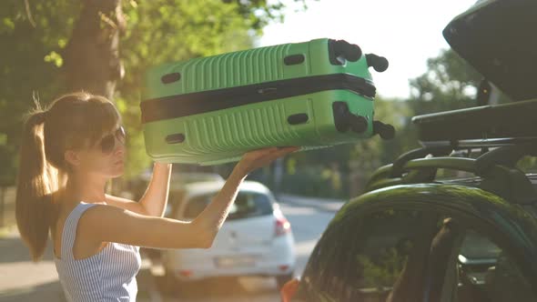 Young Woman Driver Putting Green Suitcase Inside Car Roof Rack on City Street