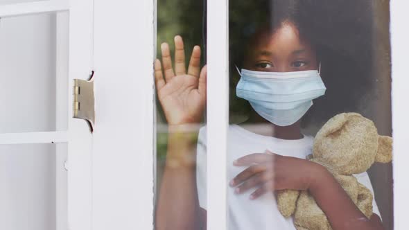 African american girl wearing face mask holding teddy bear waving through window
