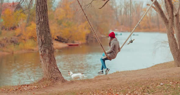 Small Dog Is Standing By the River, and the Mistress Is Swinging on a Swing. Girl Holds a Puppy