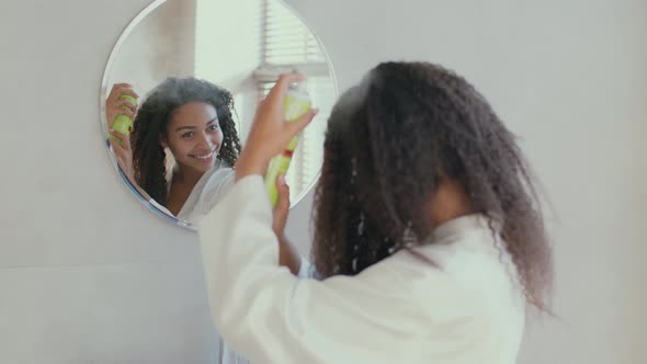 Young African American Woman Applying Hair Spray on Curly Hair Making Hairstyle at Home Looking at