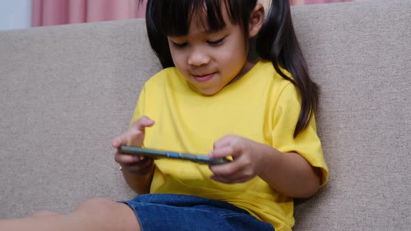 Cute little girl playing online games on her smartphone sitting on the sofa at home.