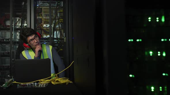 Asian male it technician with headphones on using laptop and checking computer server