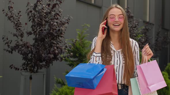 Positive Young Girl with Bags Talking on Mobile Phone About Sales in Shopping Mall in Black Friday