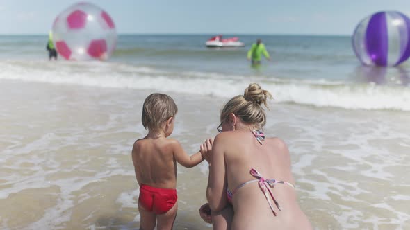 The Kid Looks at the Horizon of the Sea and Inflatable Big Balls with His Mother in a Bikini