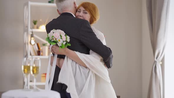 Happy Senior Bride in Wedding Dress Hugging Elegant Groom in Suit Looking at Camera Smiling