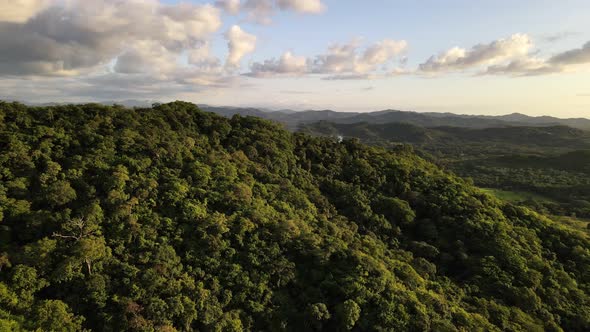 Aerial view of approach towards a mountain ridge covered by thick tropical rainforest during sunset.