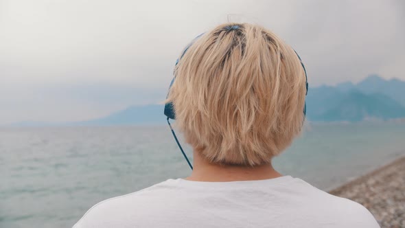 Blonde Man Sits on the Shore and Puts on the Headphones on His Head