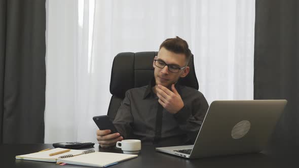 Young successful businessman in glasses looking on phone sitting at his office desk