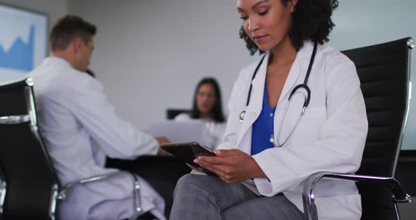 Mixed race female doctor sitting in meeting room using tablet looking to camera smiling