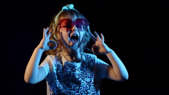 Portrait of Young Blond Girl in Bright Colorful Neon Rays Light Showing Ok Sign on Black Background