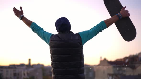 Back View Confident Man with Skateboard Stretching Hands Admiring City at Sunset on Roof