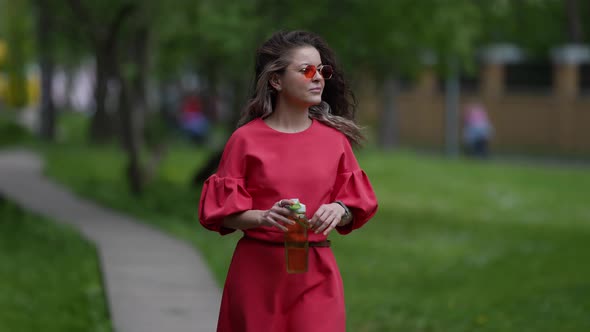 Relaxed Woman in Red Dress is Walking Alone in City Park and Drinking Water From Plastic Reusable