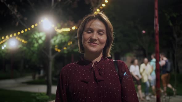 Woman smiling and looking straight to the camera on the background of evening lights of a city.