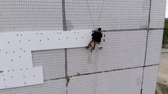 Aerial View on Industrial Climber Suspended on Ropes Performs Work on Insulation of Facade of High