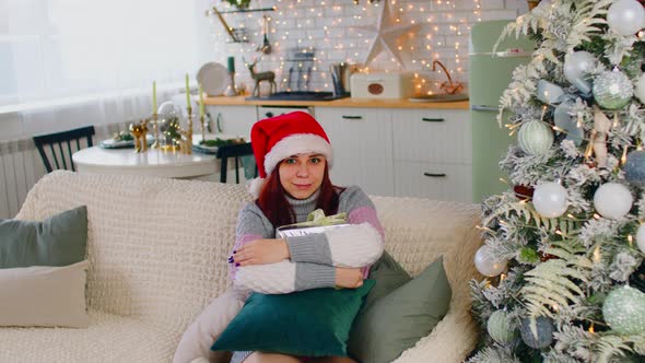 Young Woman in Santa Hat with Gift Box Sitting on Couch Near Christmas Tree in Living Room